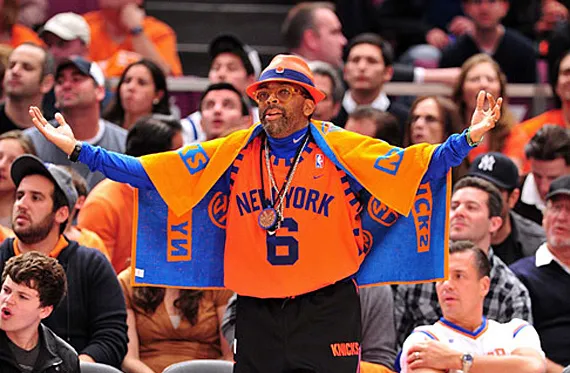 New York Knicks fan Spike Lee stands in a crowded basketball arena, wearing bright orange Knicks apparel, including a jersey with the number 6, a matching cape, and a large team towel draped over his shoulders. He holds his arms wide open, displaying team pride, surrounded by other spectators dressed in Knicks and sports attire.