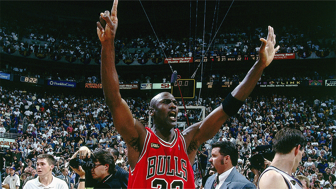 basketball player Michael Jordan in a Bulls jersey celebrates with arms raised, surrounded by an enthusiastic crowd and photographers.