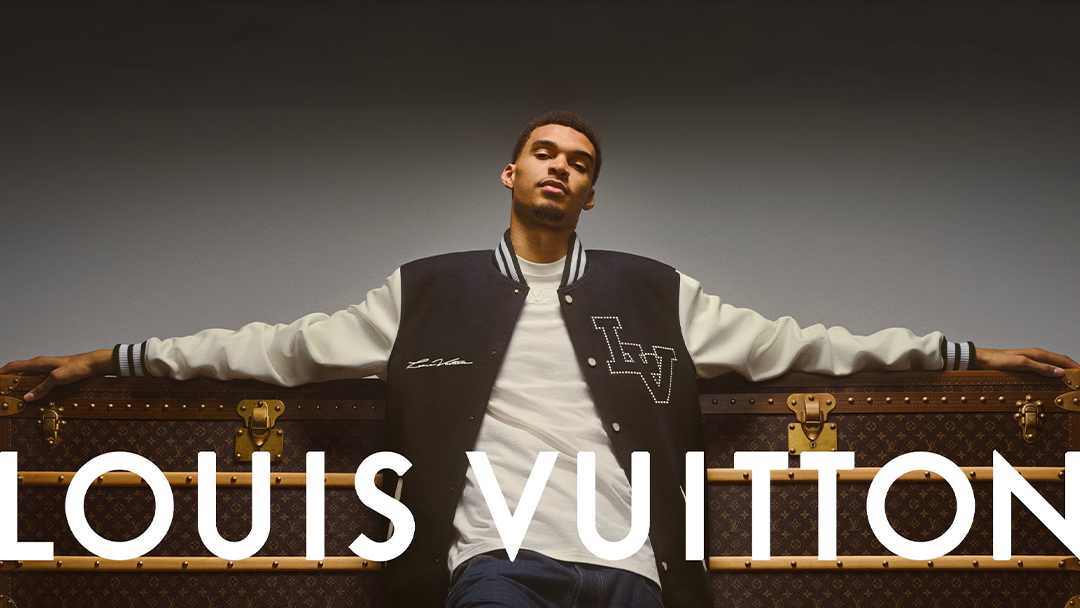 victor wembanyama wearing a black and white Louis Vuitton varsity jacket and white t-shirt sits with arms outstretched on a large Louis Vuitton monogram trunk, with bold "LOUIS VUITTON" text overlaying the image.