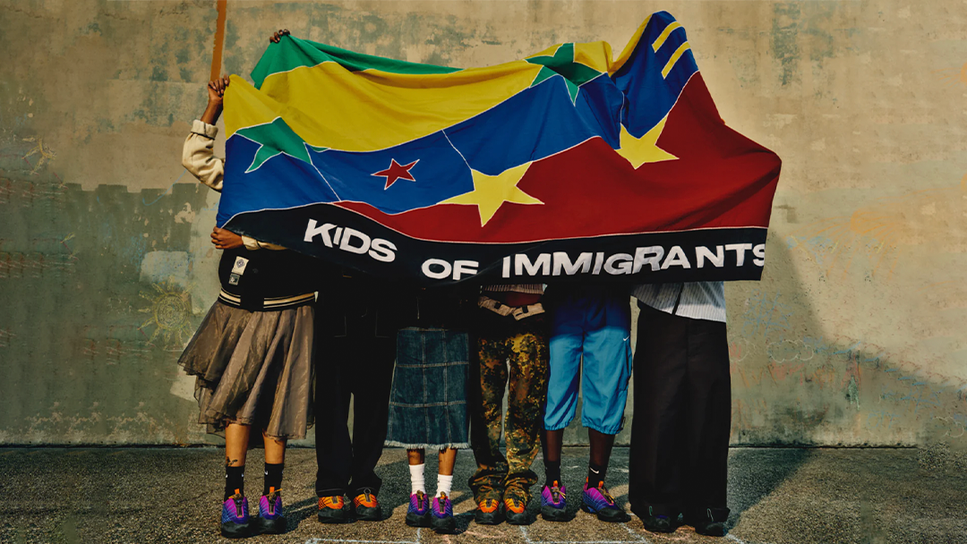 A group of people standing outdoors on the street, holding a large flag that reads "KIDS OF IMMIGRANTS." They are dressed in various clothing and footwear.