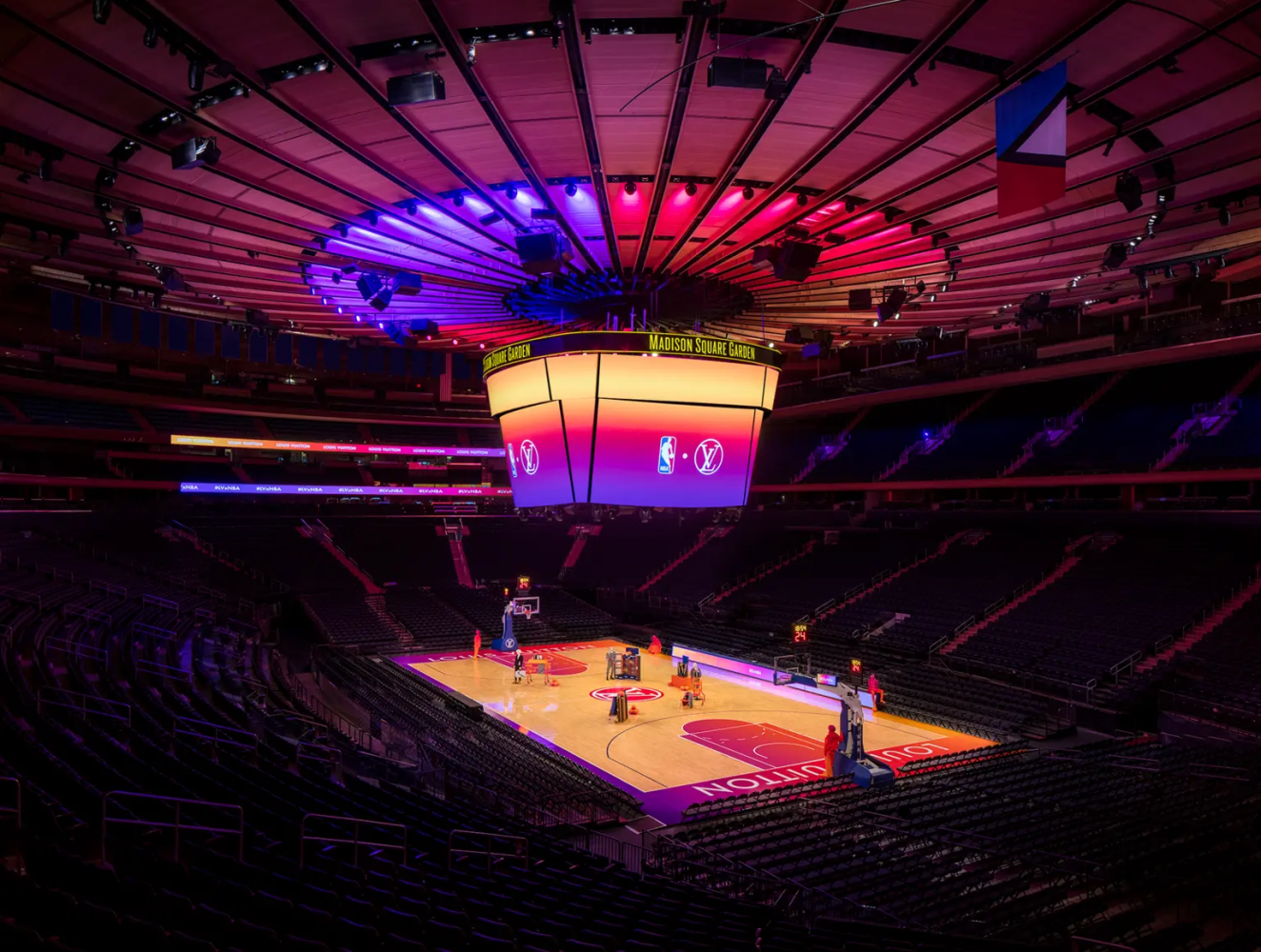 An empty Madison Square Garden basketball arena, illuminated with vibrant purple and orange lighting, showcases a brightly lit central scoreboard displaying NBA logos over the court, with no spectators in the seats.